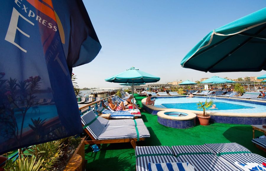 Outdoor high-top tables and lounge seating on a cruise ship deck at sunset.