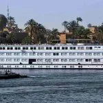 The long, white exterior of the M/S Royal Amira Nile cruise ship with multiple rows of cabin windows, sailing past a town with palm trees.