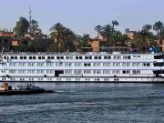 The long, white exterior of the M/S Royal Amira Nile cruise ship with multiple rows of cabin windows, sailing past a town with palm trees.