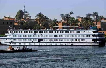 The long, white exterior of the M/S Royal Amira Nile cruise ship with multiple rows of cabin windows, sailing past a town with palm trees.