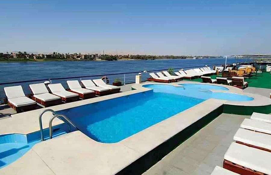 Nile cruise ship sundeck with a rectangular pool, a jacuzzi, and rows of white lounge chairs overlooking the wide blue river.
