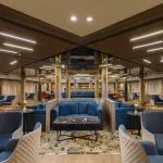 Luxurious cruise ship lounge featuring symmetrical seating areas with plush blue and cream chairs, dark wood ceilings, and reflective central pillars.