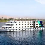 The Nile Crown cruise ship, a large white vessel with blue and turquoise geometric accents, sailing on the clear blue Nile River with palm trees and arid hills visible on the far bank under a bright sky.