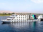 The Nile Crown cruise ship, a large white vessel with blue and turquoise geometric accents, sailing on the clear blue Nile River with palm trees and arid hills visible on the far bank under a bright sky.