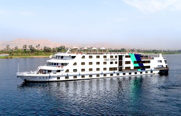 The Nile Crown cruise ship, a large white vessel with blue and turquoise geometric accents, sailing on the clear blue Nile River with palm trees and arid hills visible on the far bank under a bright sky.