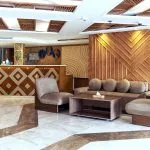 The modern reception lobby of a cruise ship, featuring a distinctive wooden check-in desk with diamond patterning, a seating area with taupe chairs and a sofa, and patterned marble flooring, all accented by decorative wood wall panels.