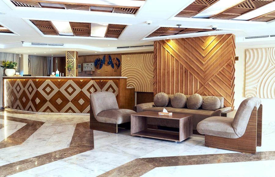 The modern reception lobby of a cruise ship, featuring a distinctive wooden check-in desk with diamond patterning, a seating area with taupe chairs and a sofa, and patterned marble flooring, all accented by decorative wood wall panels.