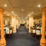 The interior of a cruise ship dining room, characterized by rows of large, orange-hued decorative columns, a dark blue carpet, and multiple round dining tables set with light blue tablecloths.