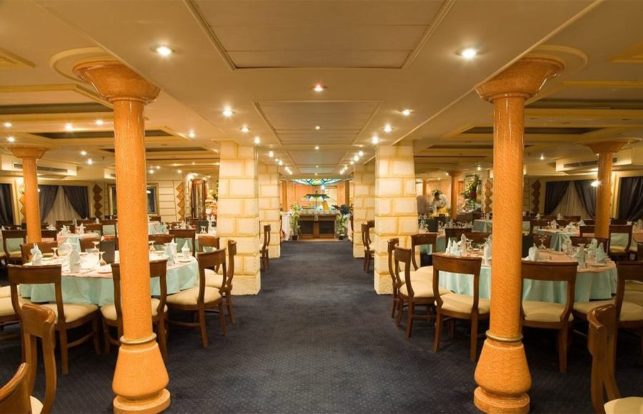 The interior of a cruise ship dining room, characterized by rows of large, orange-hued decorative columns, a dark blue carpet, and multiple round dining tables set with light blue tablecloths.