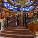 A lavish cruise ship atrium featuring a grand, curved staircase with ornate black wrought-iron railings, rich wood paneling, and a large, colorful circular stained-glass dome ceiling above.