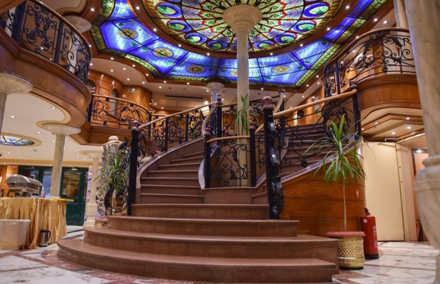 A lavish cruise ship atrium featuring a grand, curved staircase with ornate black wrought-iron railings, rich wood paneling, and a large, colorful circular stained-glass dome ceiling above.