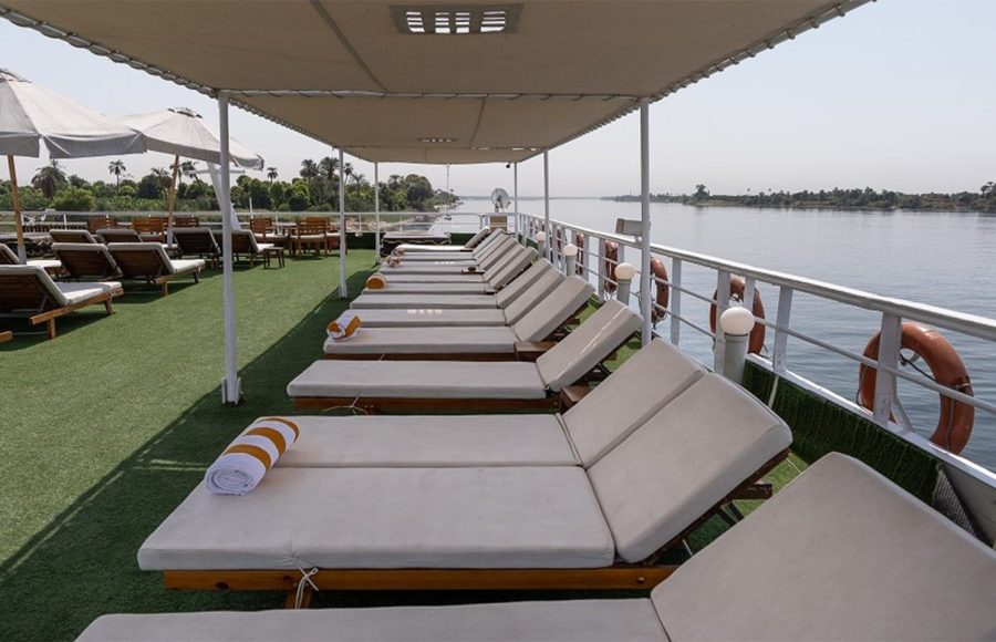 A row of wooden sun loungers with thick, white cushions and rolled orange-striped towels on a cruise ship sundeck with green artificial grass, facing the Nile River and the lush, palm-dotted shoreline.