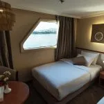 Close-up view inside a cruise ship twin cabin showing one of the beds, a cozy armchair, and an irregularly shaped window offering a clear view of the Nile River and distant mountains under a bright sky.