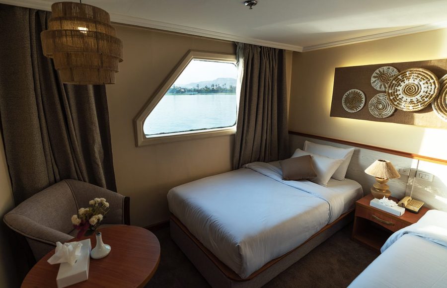 Close-up view inside a cruise ship twin cabin showing one of the beds, a cozy armchair, and an irregularly shaped window offering a clear view of the Nile River and distant mountains under a bright sky.