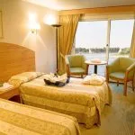 A brightly lit twin cabin on a Nile cruise ship featuring two single beds with patterned gold and beige bedspreads, a sitting area with two green armchairs by a large window overlooking the river, and a small television.