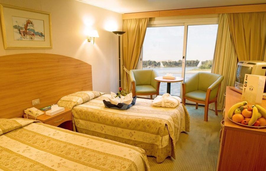 A brightly lit twin cabin on a Nile cruise ship featuring two single beds with patterned gold and beige bedspreads, a sitting area with two green armchairs by a large window overlooking the river, and a small television.