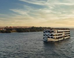 A multi deck standard Nile cruise ship sailing on the wide, calm Nile River with green banks and a clear sky, representing a comfortable and scenic way to experience the best standard Nile cruises.
