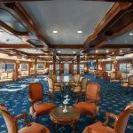 Spacious cruise ship lounge with dark wood paneling, a blue patterned carpet, and multiple conversational seating arrangements.