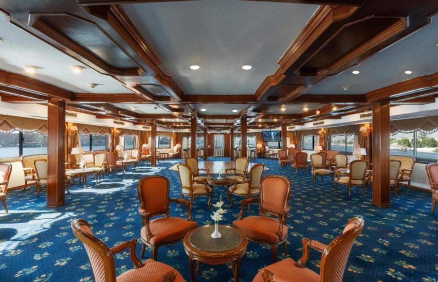 Spacious cruise ship lounge with dark wood paneling, a blue patterned carpet, and multiple conversational seating arrangements.
