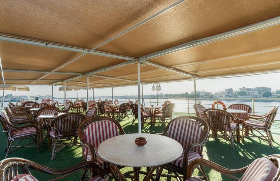 Shaded outdoor seating area on a cruise ship sundeck with many rattan chairs and red/white striped cushions, overlooking the Nile.