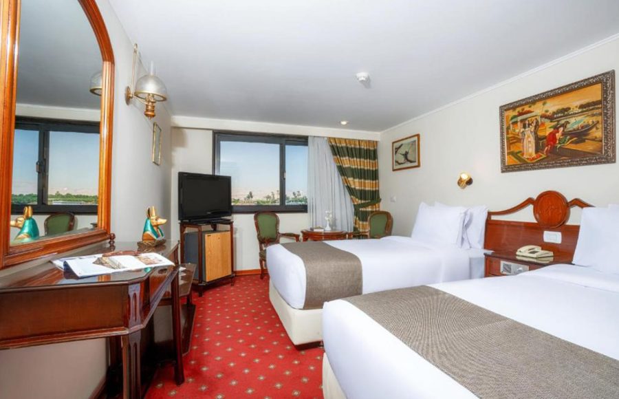 Traditional-style twin cabin on a Nile cruise with two beds, red patterned carpet, dark wood furniture, and a window view of the river.