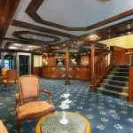 Traditional-style cruise ship lobby with a wooden reception desk, a carpeted staircase, and classic seating areas.