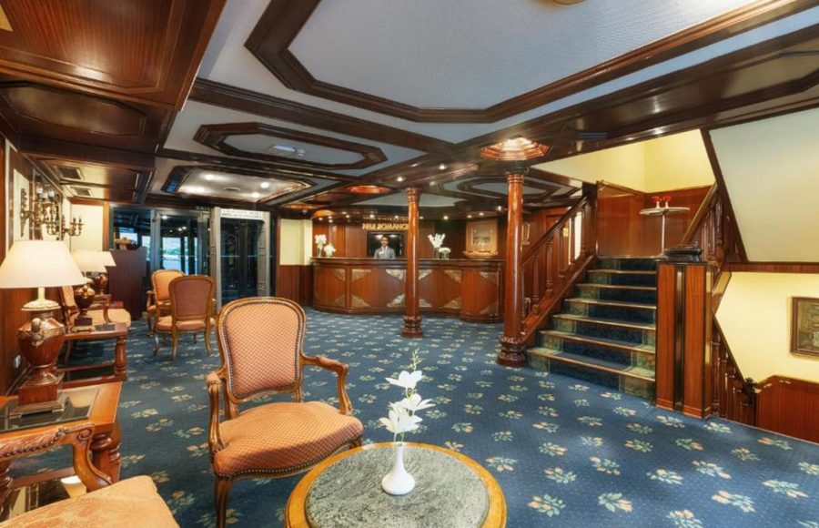 Traditional-style cruise ship lobby with a wooden reception desk, a carpeted staircase, and classic seating areas.