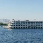 A wide, white, multi-deck river cruise ship named "Nile Romance" sailing on the blue river under a clear sky.