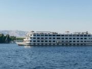 A wide, white, multi-deck river cruise ship named "Nile Romance" sailing on the blue river under a clear sky.
