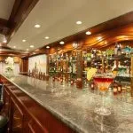 Interior view of a cruise ship lobby lounge with multiple beige couches, wood-paneled columns, and a bar in the background.