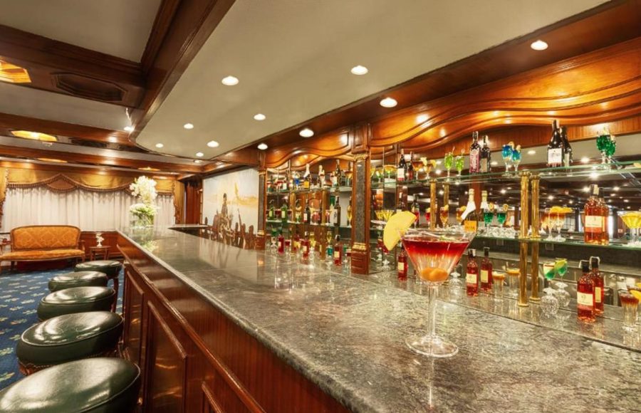 Interior view of a cruise ship lobby lounge with multiple beige couches, wood-paneled columns, and a bar in the background.