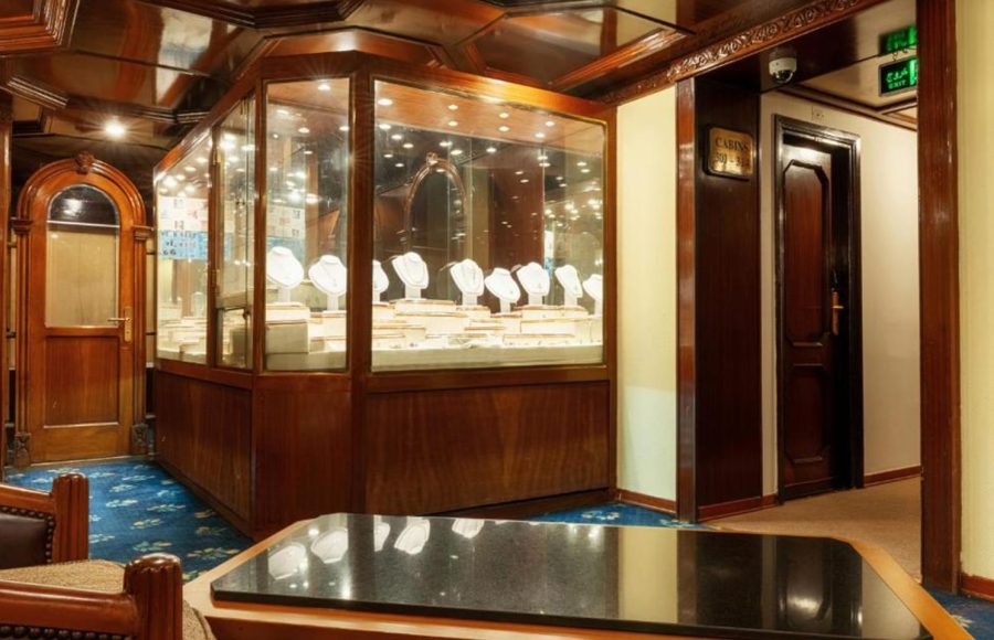 Interior view of a cruise ship jewelry shop with a large, illuminated wooden display case showing necklaces and pendants.
