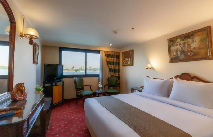 Traditional-style Nile cruise cabin with a large king bed, red patterned carpet, and view of the river from the window.