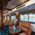 Traditional-style cruise ship lounge with dark wood trim, classic upholstered furniture, and large windows overlooking the river.