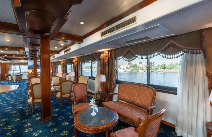 Traditional-style cruise ship lounge with dark wood trim, classic upholstered furniture, and large windows overlooking the river.
