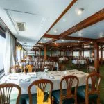 Wide view of a cruise ship dining room with dark wooden chairs, tables with white linens, and large panoramic windows overlooking the Nile.