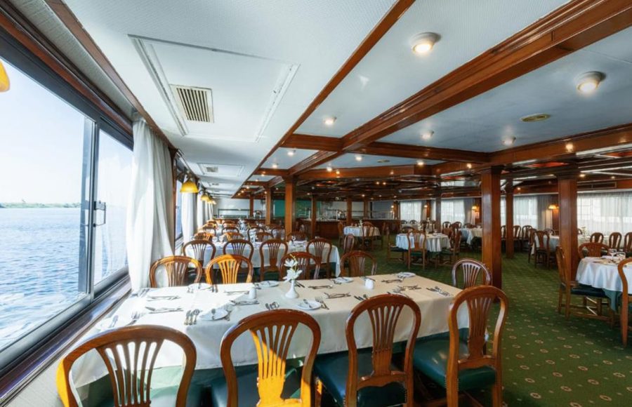Wide view of a cruise ship dining room with dark wooden chairs, tables with white linens, and large panoramic windows overlooking the Nile.