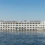 A wide, white, multi-deck river cruise ship named "Nile Romance" sailing on the blue river under a clear sky.