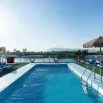 Rectangular swimming pool on a cruise ship sundeck, surrounded by lounge chairs on green turf, with a river and mountains view.