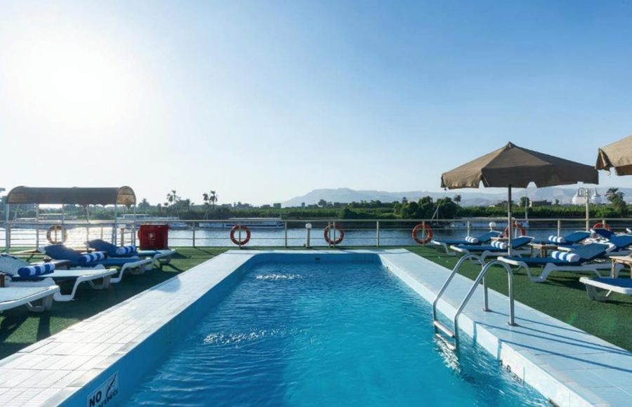 Rectangular swimming pool on a cruise ship sundeck, surrounded by lounge chairs on green turf, with a river and mountains view.