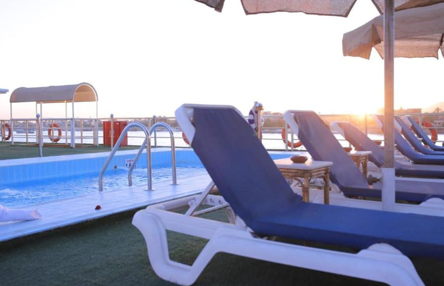 Rows of white outdoor tables and chairs under a brown canopy on the cruise ship's sun deck, overlooking the Nile River.
