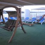 A wooden outdoor porch swing/hammock on the ship's sundeck next to blue lounge chairs and a view of the river.