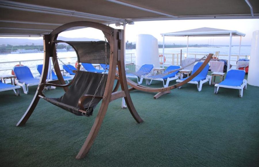 A wooden outdoor porch swing/hammock on the ship's sundeck next to blue lounge chairs and a view of the river.