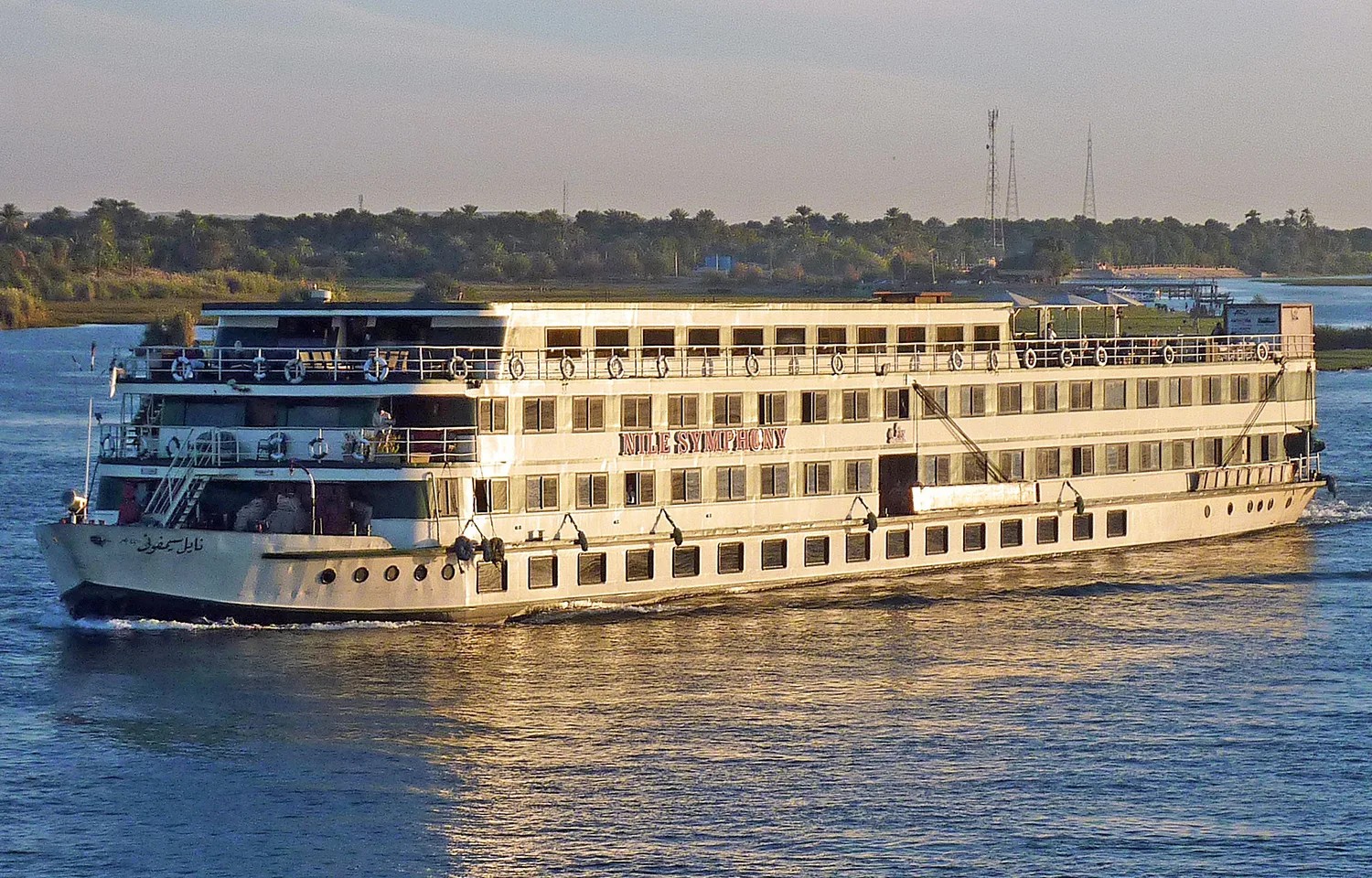 The MS Nile Symphony cruise ship sailing on the river, showing its white hull, multiple decks of cabins with windows, and the ship's name prominently displayed.