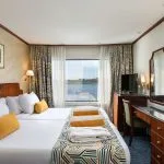A modern, brightly lit twin-bedded cabin on a Nile cruise ship with dark wood furniture, white linens, yellow accent pillows and throws, patterned curtains, and a large window overlooking the river.