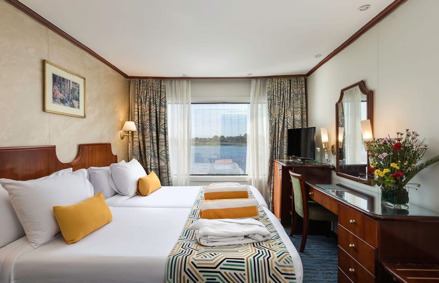 A modern, brightly lit twin-bedded cabin on a Nile cruise ship with dark wood furniture, white linens, yellow accent pillows and throws, patterned curtains, and a large window overlooking the river.
