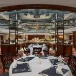 A formal cruise ship dining room (restaurant) featuring multiple round tables with white linens, elegant dark wood chairs, a red patterned carpet, and a central buffet area with a distinctive illuminated ceiling structure.