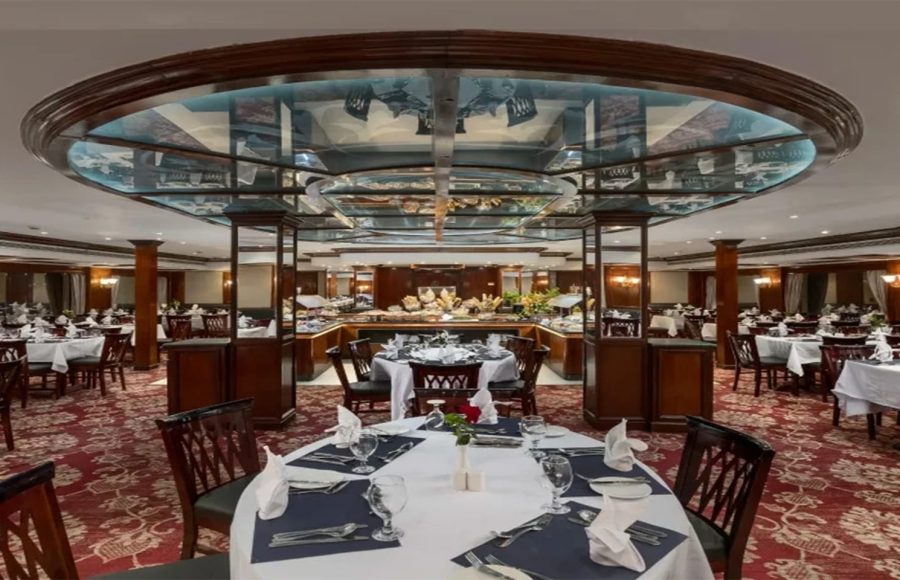 A formal cruise ship dining room (restaurant) featuring multiple round tables with white linens, elegant dark wood chairs, a red patterned carpet, and a central buffet area with a distinctive illuminated ceiling structure.