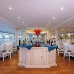 Wide view of a bright cruise ship dining room with white chairs and a central wine display.