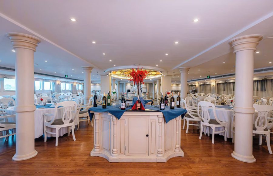 Wide view of a bright cruise ship dining room with white chairs and a central wine display.